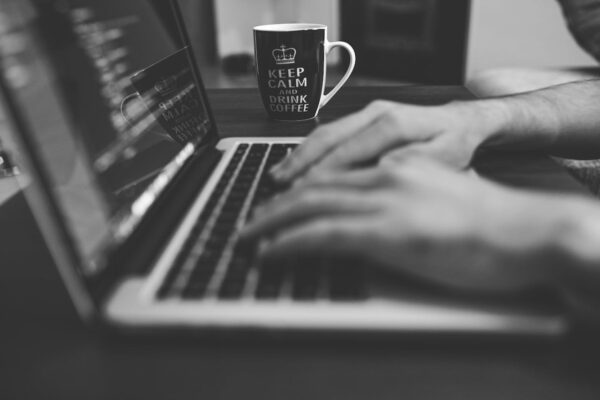 Monochrome image of hands on a laptop keyboard with a coffee mug, coding in progress.